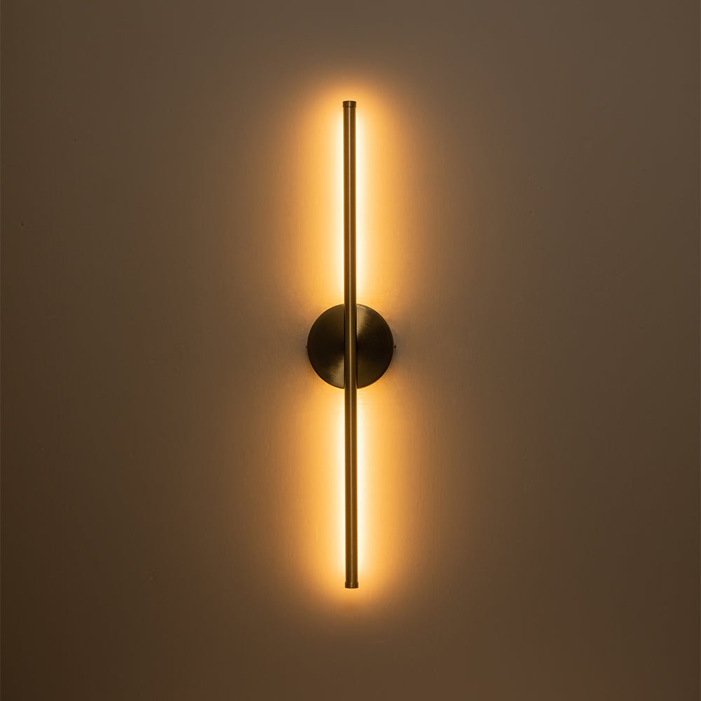 Sebastian linear LED wall light – in brass, lit on a brown wall